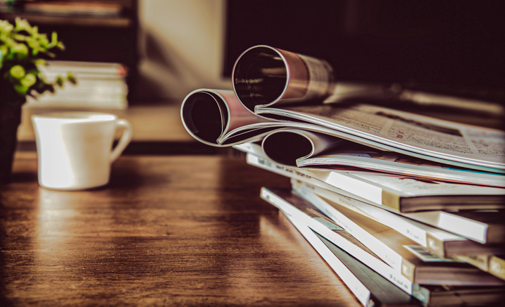 A cup of coffee and a stack of books and magazines on a wood table.