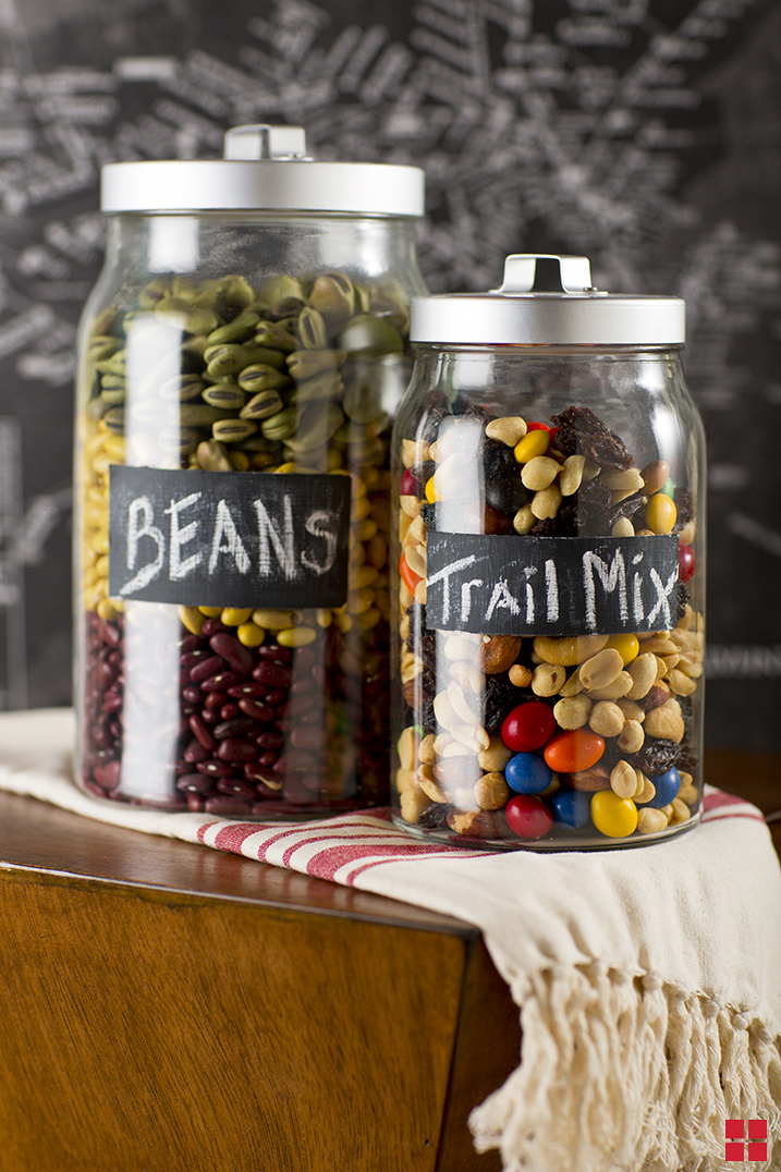 A large jar containing three kinds of dry beans with a chalkboard label that reads “Beans,” and a smaller jar filled with nuts, raisins and chocolate candy with a chalkboard label that reads “Trail Mix.” 