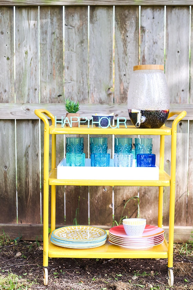 A three-tier yellow metal cart on wheels against a wooden fence. A beverage dispenser, plates, glasses and a Happy Hour sign are on the cart’s shelves.