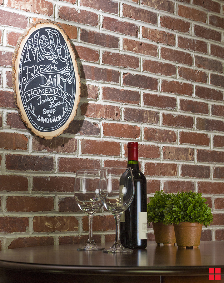 A wood slice chalkboard with a menu written on it in white chalk. The chalkboard hangs on a brick wall above a table with two glasses of wine and a wine bottle.