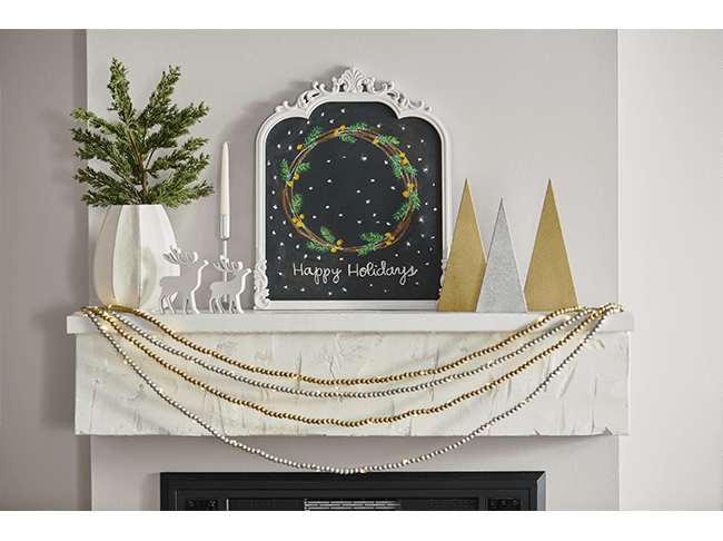 A chalkboard in a white frame on the center of a mantel. The chalkboard features a drawing of a wreath and the words Happy Holidays. 