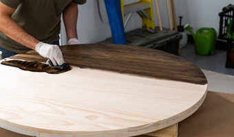 A man with a glove on his hand using a rag to apply wood stain to a bare wood table.