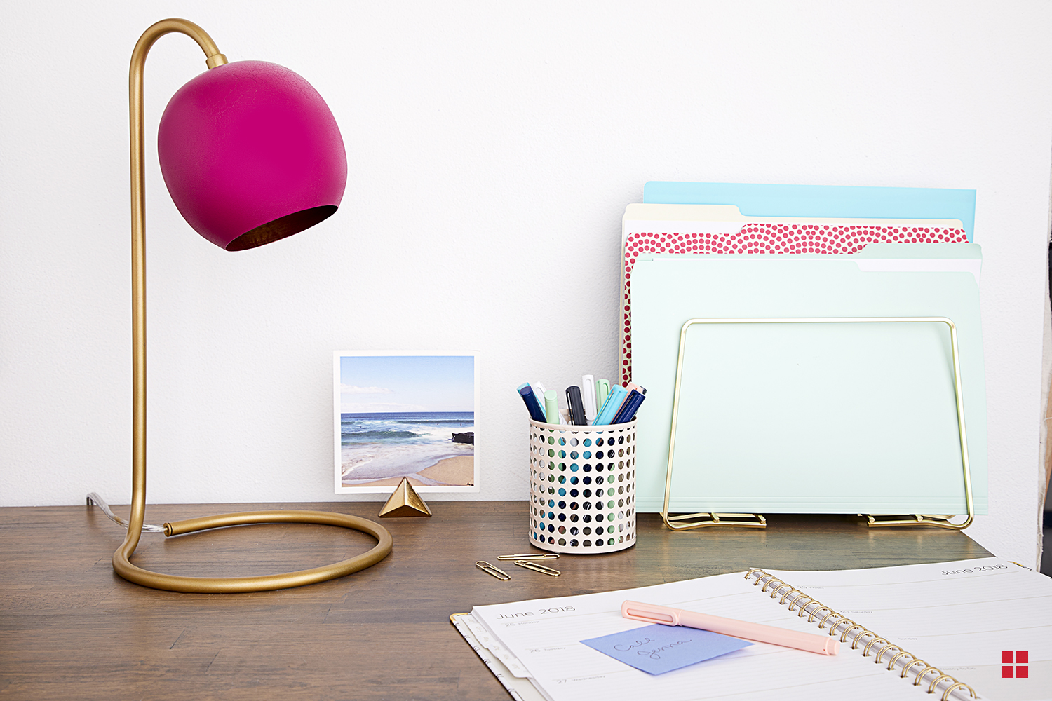 A desk with a calendar, file folders, jar of pens and a lamp with a gold base and magenta shade.