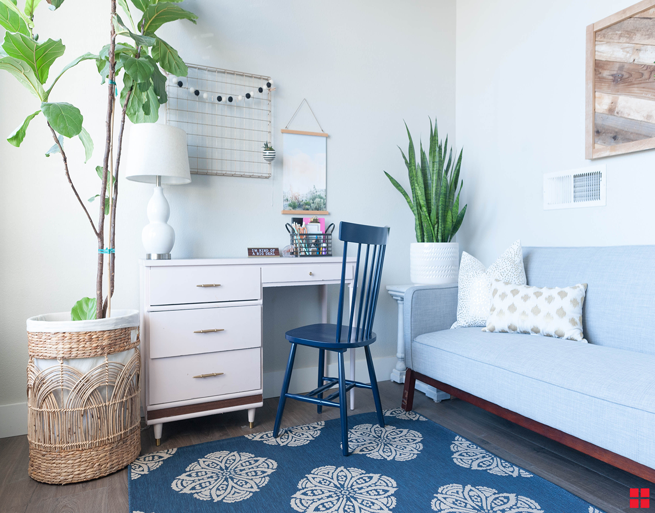 A deep blue wooden chair in front of a white desk. Potted plants are on either side of the desk, and a light blue couch is on an adjacent wall. 
