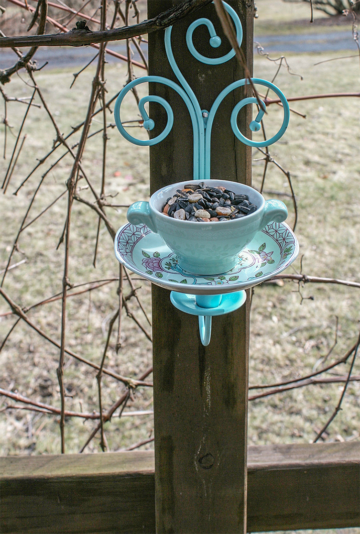 Bird feeder made out of a ceramic tea cup and saucer held in place with artfully crafted metal rods attached to a piece of wood