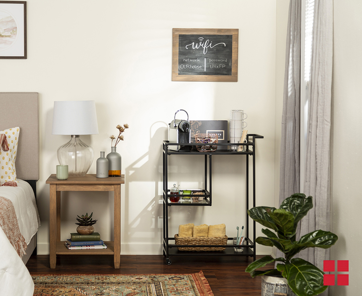 A black cart next to a wooden nightstand in a guest bedroom. The cart has three shelves which carry coffee cups, towels and a cup with toothbrushes in it. 