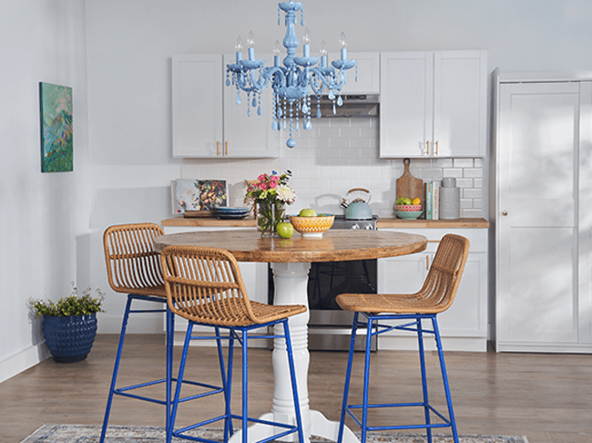 Three wooden stools with dark blue legs arranged around a kitchen table with a light blue chandelier. 