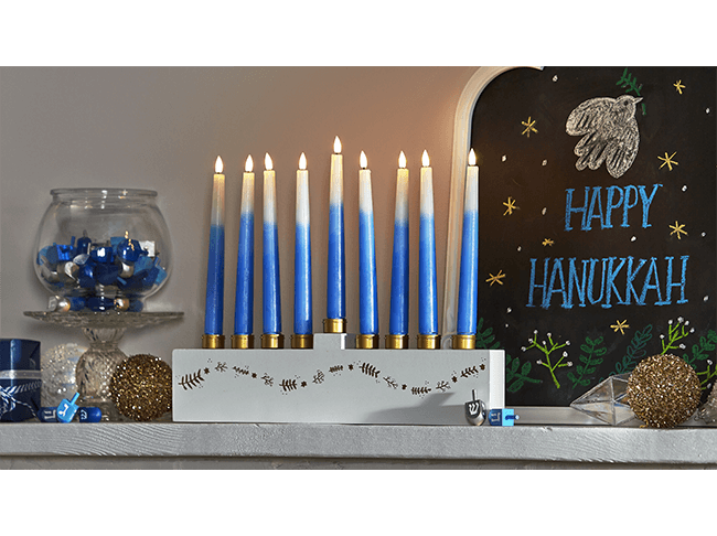 A white and gold menorah with blue and white candles displayed on a mantel next to a Happy Hanukkah sign. 