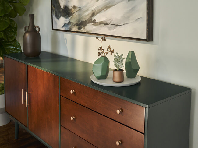 Dining room buffet painted dark green with natural wood doors and copper hardware with a gray charger plate and green and copper vases.
