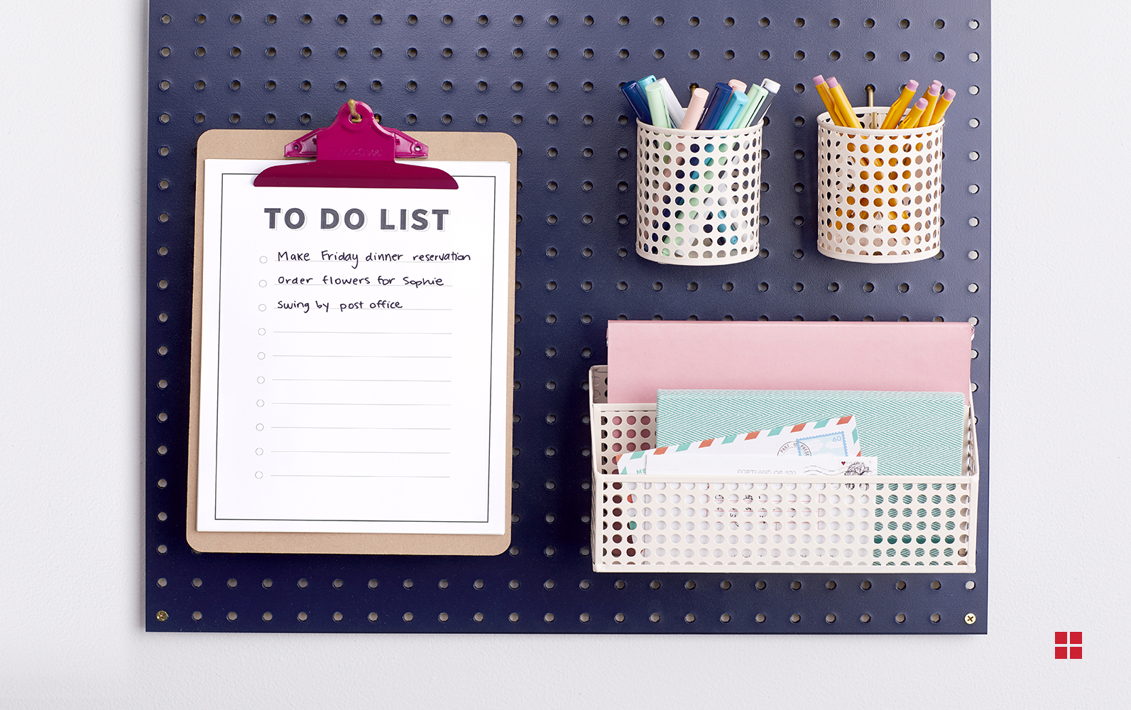 A navy blue pegboard holding a clipboard with a to-do list, two cups full of pens and pencils and a caddy containing paper and envelopes.