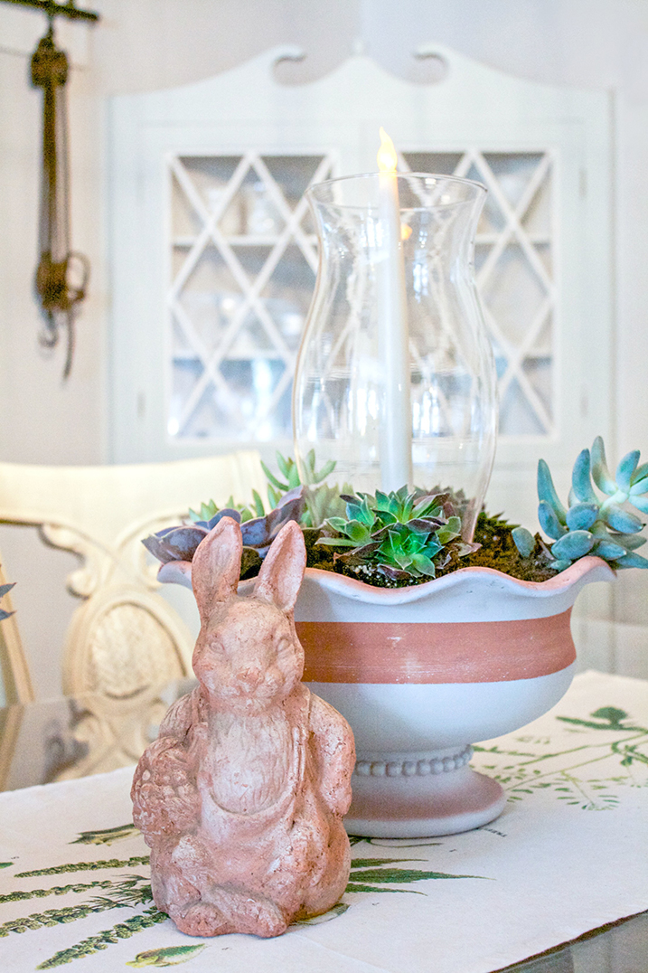 Painted flower pot with a plant and candle in it with a bunny ornament positioned next to it