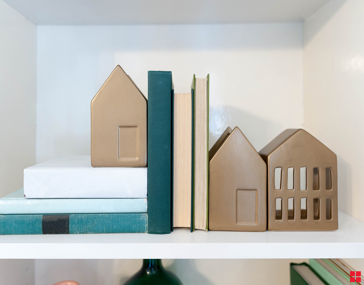 Three metallic gold bookends shaped like houses on a shelf with books. 