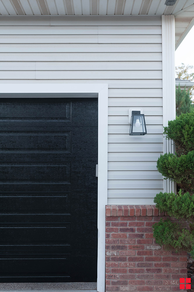 The side of a black garage door on a garage with gray siding and brick below it.