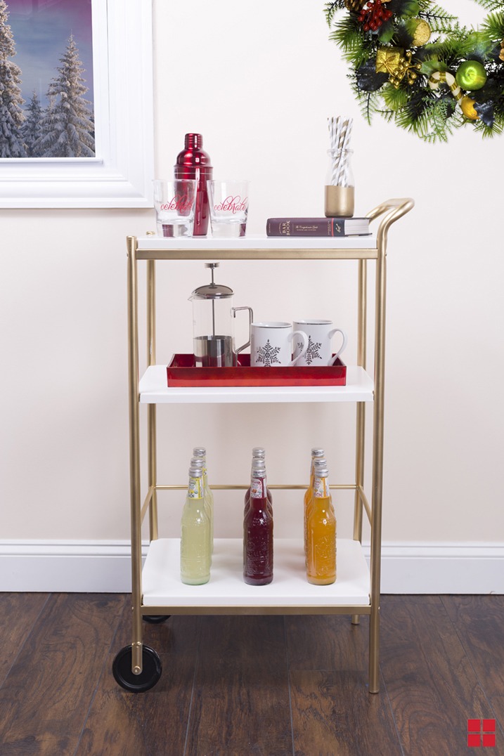 A gold bar cart with white shelves holding glasses, mugs and bottled beverages. 