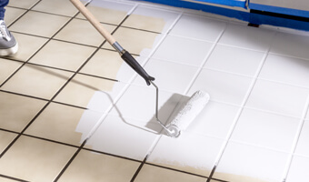 Using a paint roller to apply white tile paint over a tan tile floor. 