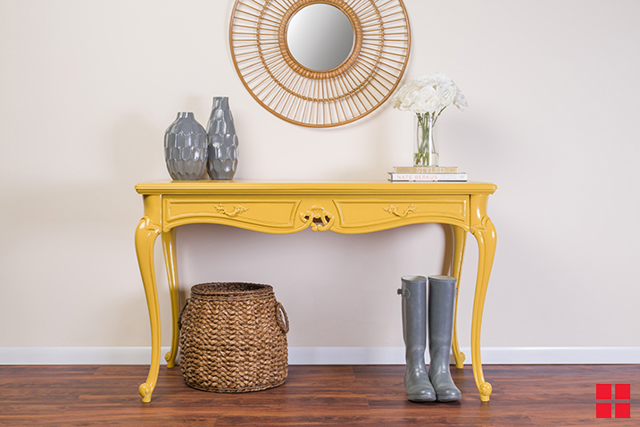 A yellow table with vases on it against a beige wall. A mirror hangs over the table and a wicker basket and pair of rain boots are below it. 