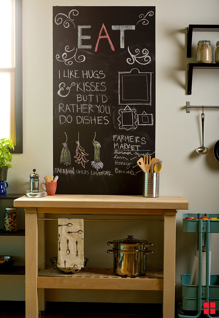 A chalkboard wall above a kitchen storage unit. The chalkboard says “Eat,” “I like hugs and kisses but I’d rather you do the dishes” and has drawings of herbs on it. 