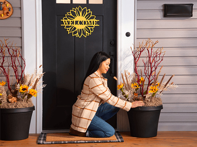 A woman kneeling on porch next to an urn planter full of fall foliage. 