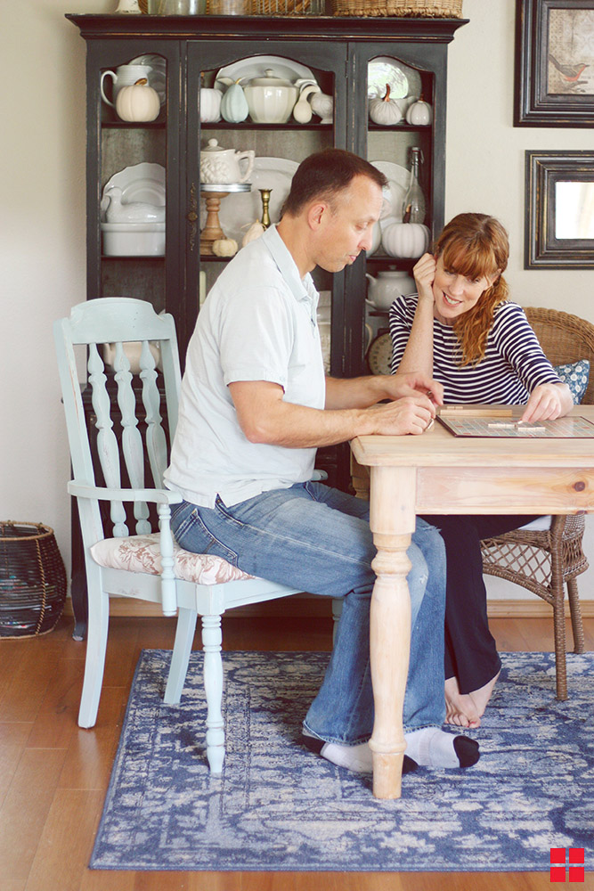 Man sitting on a pale blue chair while playing Scrabble with a woman. 
