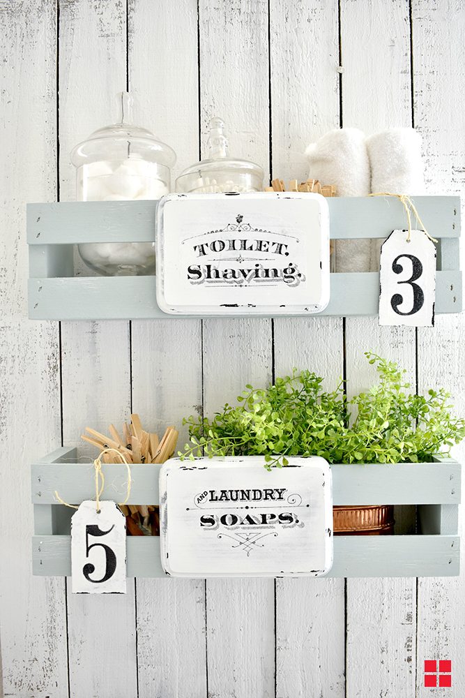 A white shiplap wall with two pale blue wooden shelves hanging on it. The top shelf is labeled toilet, shaving, and the bottom shelf is labeled laundry soaps. 