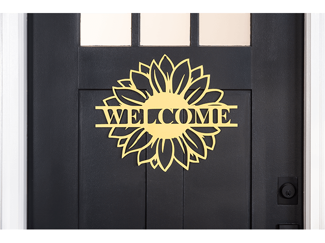 A black front door with a yellow sign shaped like a sunflower and the word “welcome” in the center. 